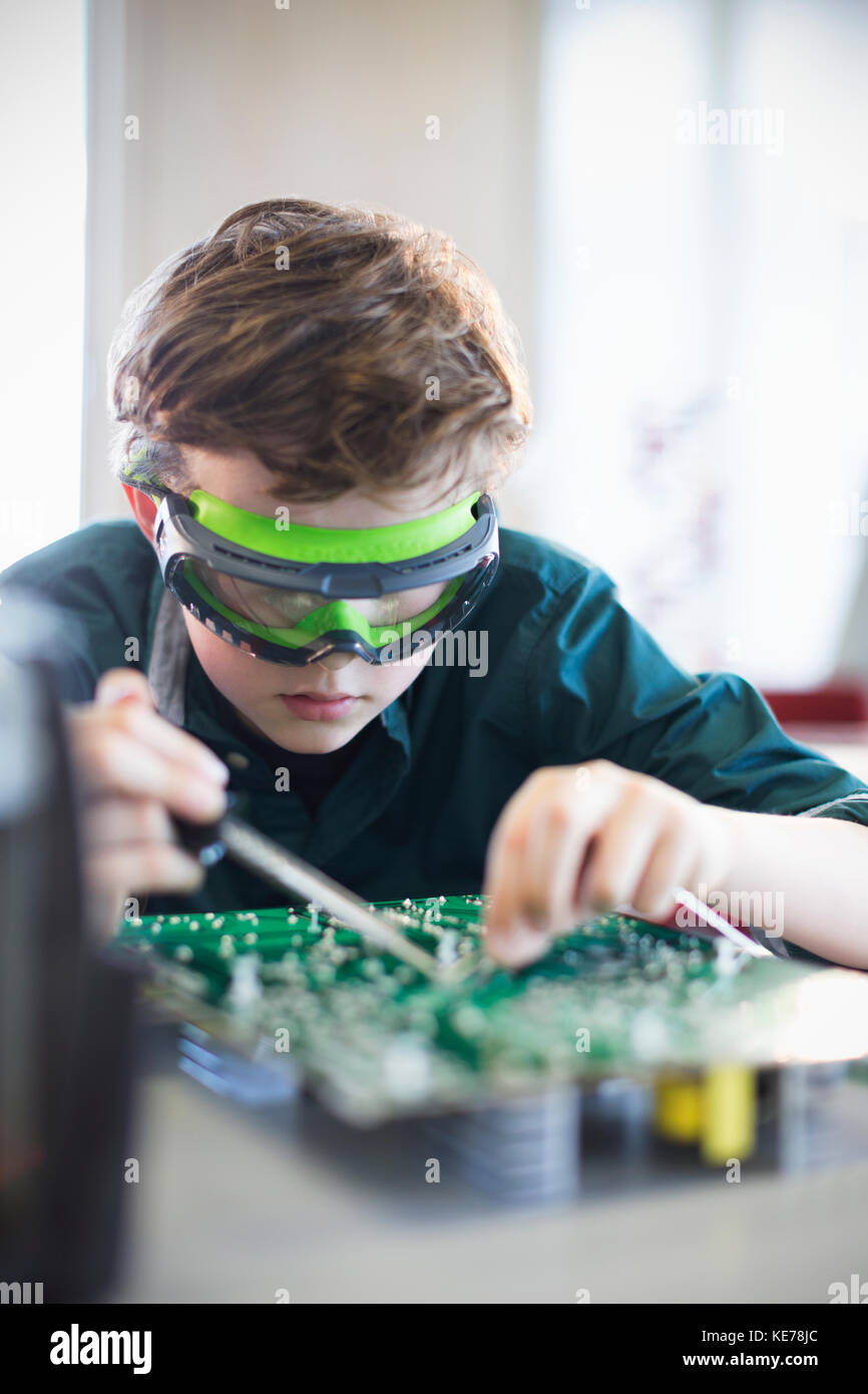 Focused boy student in goggles soldering circuit board in classroom