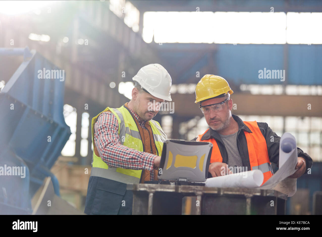 Male foreman and engineer working at laptop with blueprints in factory Stock Photo