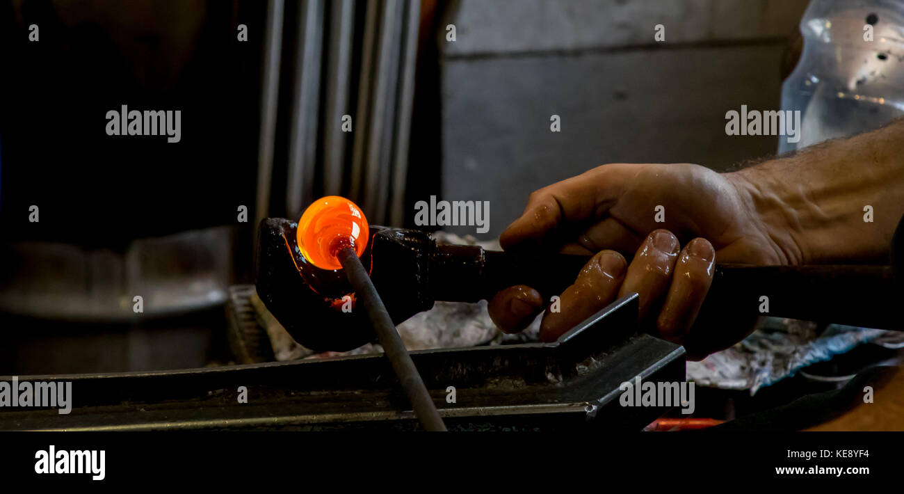 Close up of traditional craftsman at work in glassblowing studio. Man's hand gripping tools, shaping glowing molten glass. UK glassblowing art. Stock Photo
