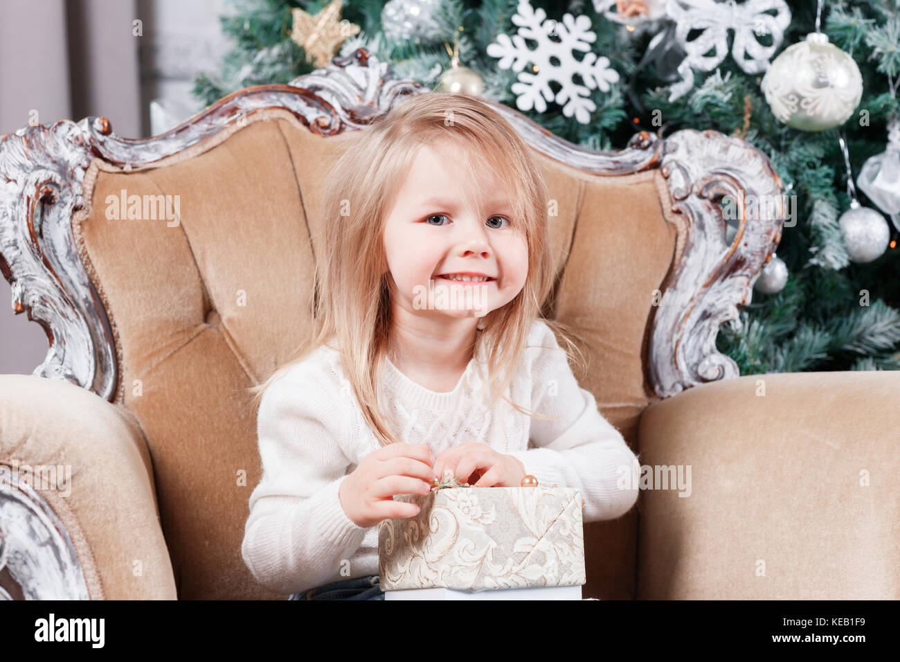 cute little girl sitting in a chair and opens a box with a present for ...