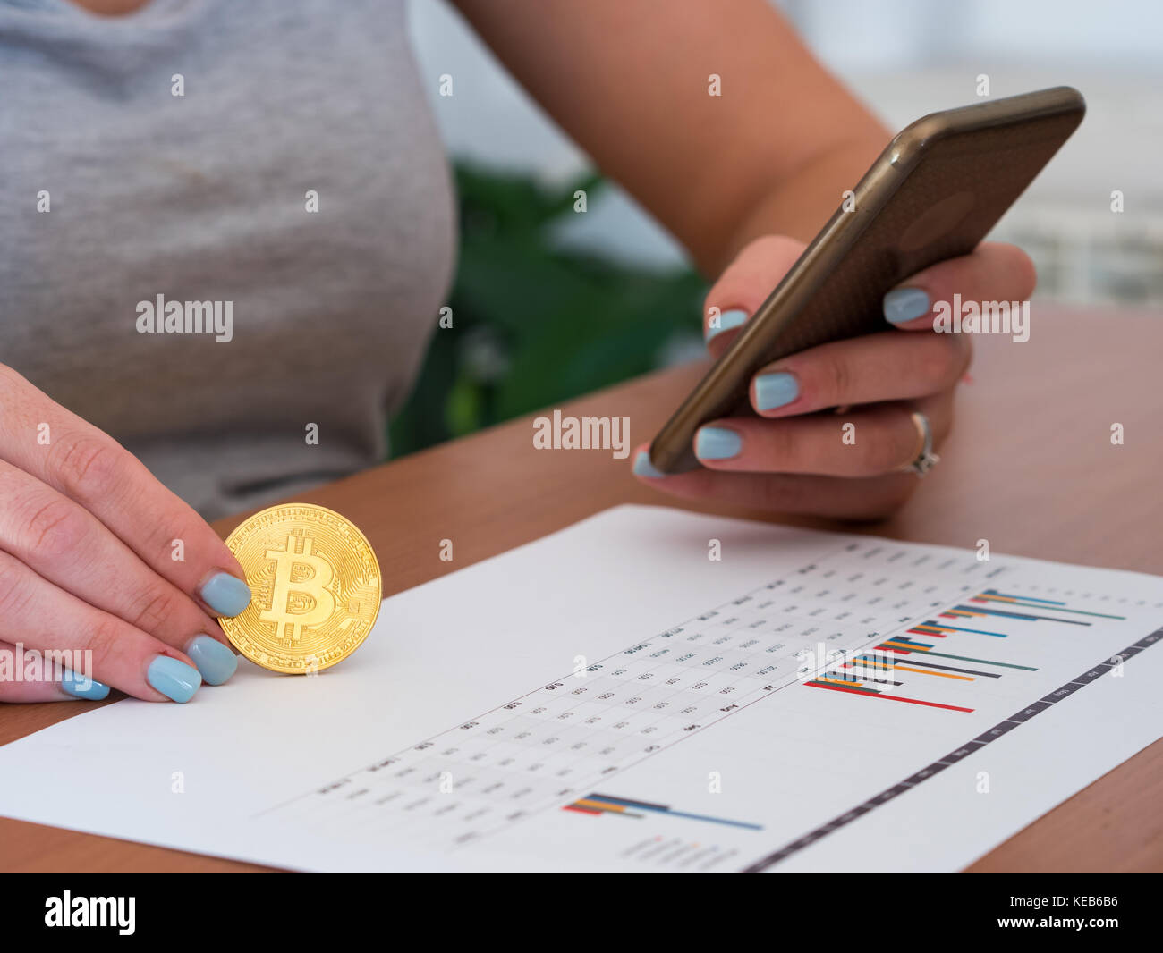 Young caucasian woman holding bitcoin on office desk and typing on smartphone Stock Photo