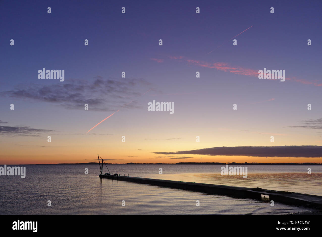 Pier before sunrise with small crane silhouetted against the morning sky and bright red vapor trail overhead Stock Photo