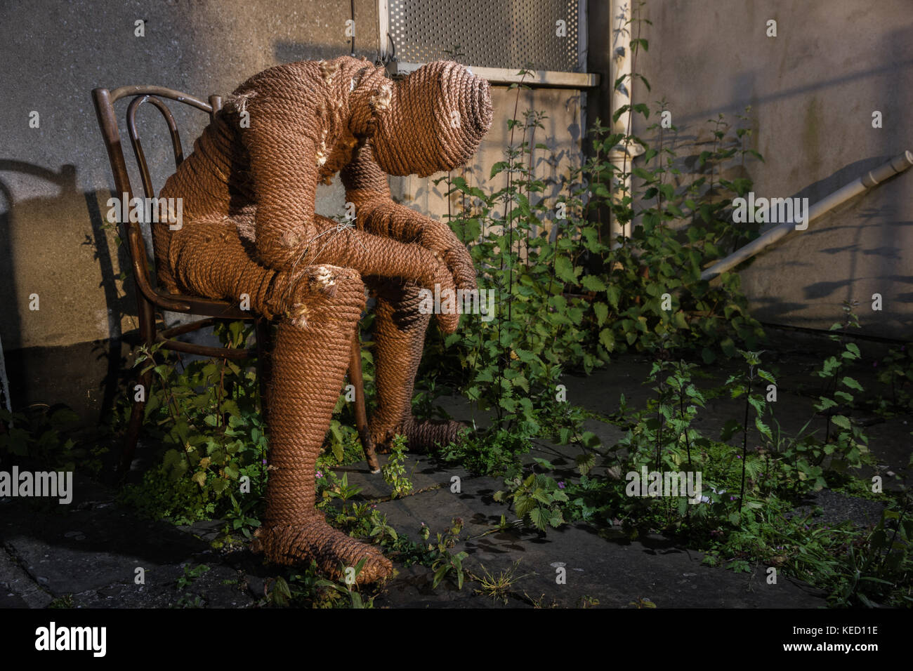 A figure made of rope sitting on a chair in a weedy concrete garden. Stock Photo