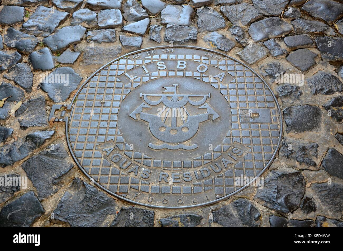 An old manhole cover in  a cobbled street in Lisbon with a sailing ship symbol and the word Lisboa Portugal Stock Photo