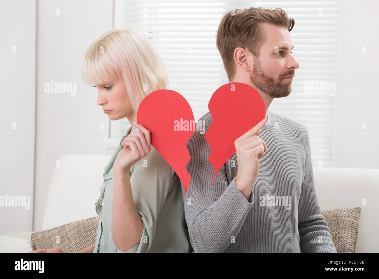 Sad Couple Sitting On Sofa Holding Red Broken Heart At Home Stock Photo