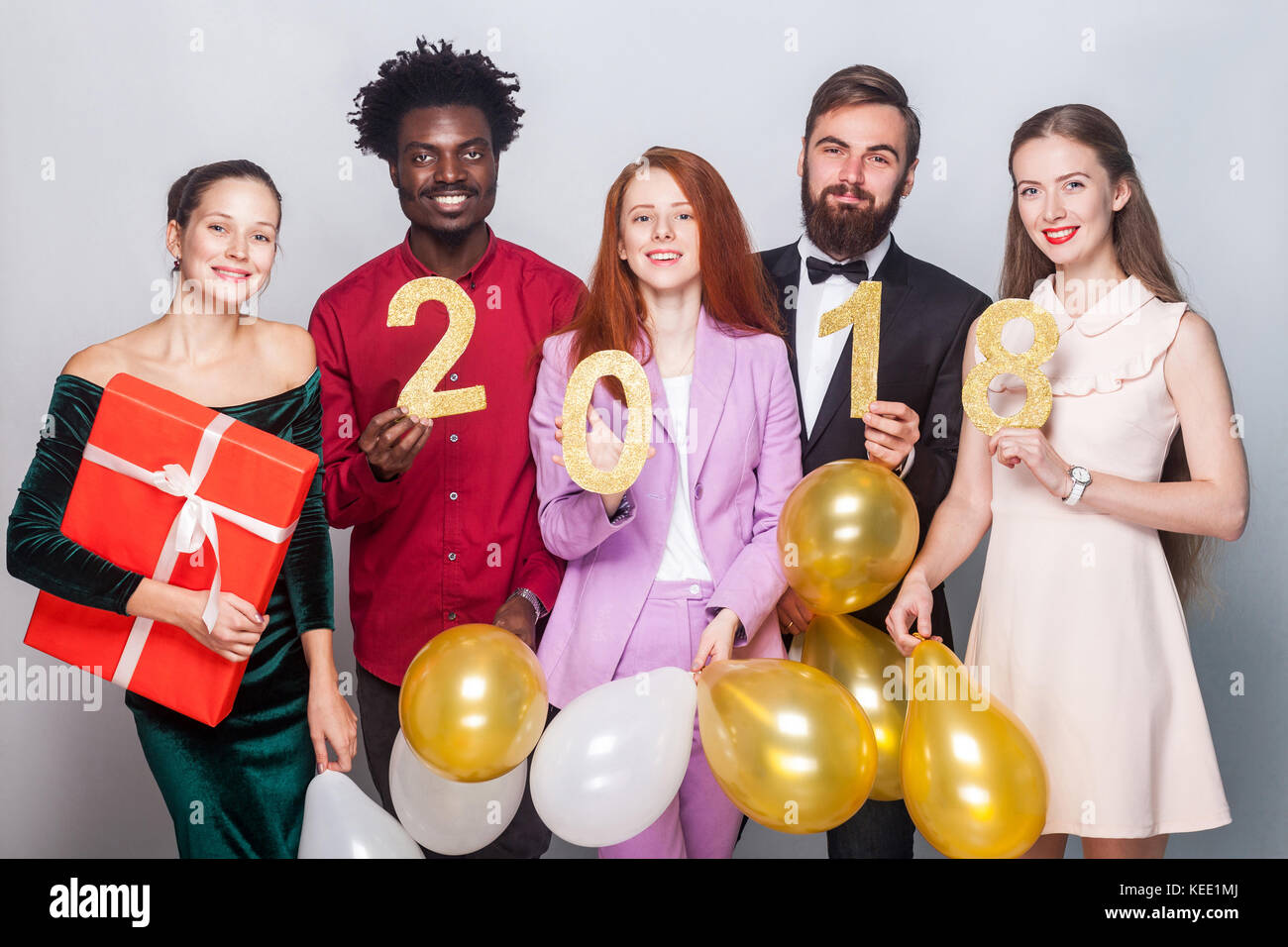 Well dressed beautiful people celebrating new year. Holding gift box, colorful air balloon and number 2018, looking at camera and smiling. Studio shot Stock Photo