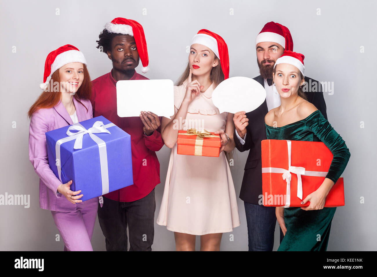 Beautiful long haired blonde girl looking at camera with puzzlement and dreaming face, her friends standing near and holding gift box. Studio shot. Gr Stock Photo