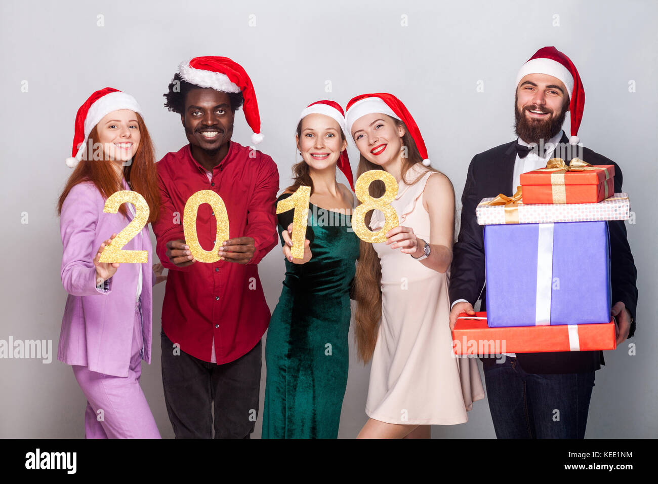 Young adult best friends holding number 2018  and looking at camera with toothy smiling. One bearded man standing near and holding many gift box.  Ind Stock Photo