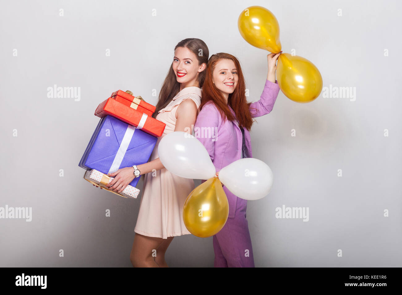 Beautiful girls standing back to back, holding many box and air balloon, toothy smiling and looking at camera. Studio shot, gray background Stock Photo