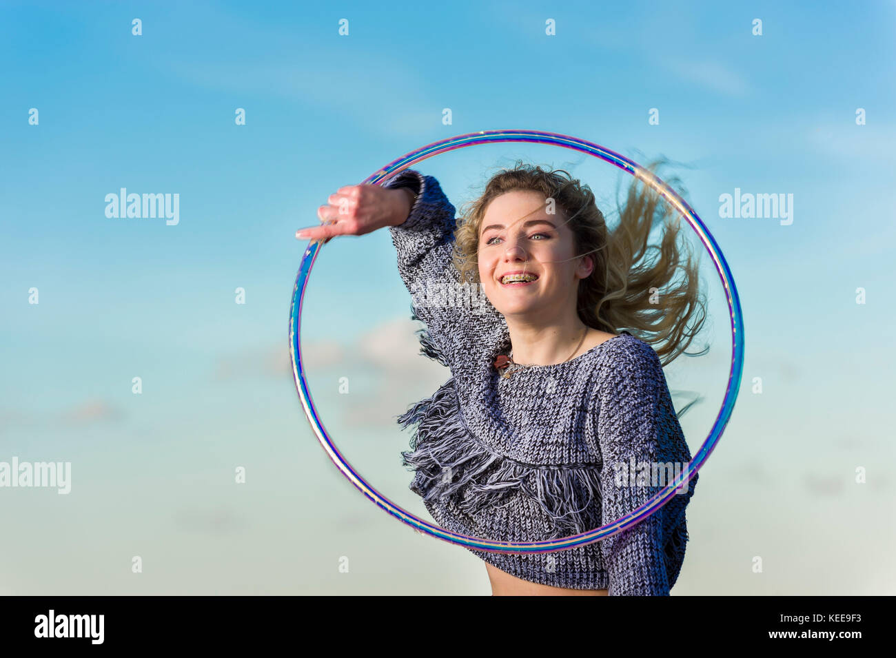 Girl smiles as she performs hooping, captured in mid air, looking through hoop against a blue sky with some small clouds. Stock Photo