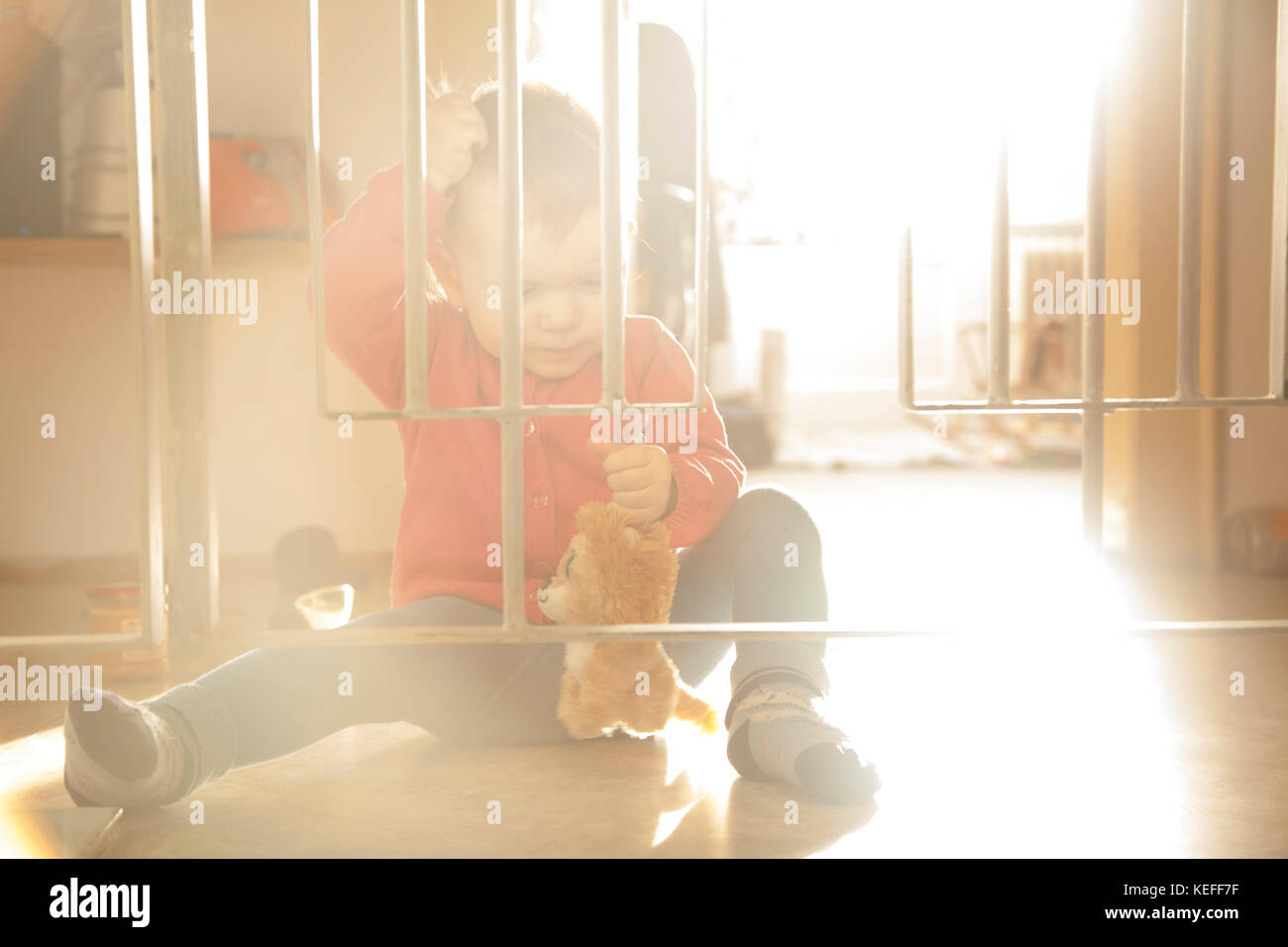 Little baby girl playing with toy behind the bars inside of the house in the morning sunlight. Stock Photo