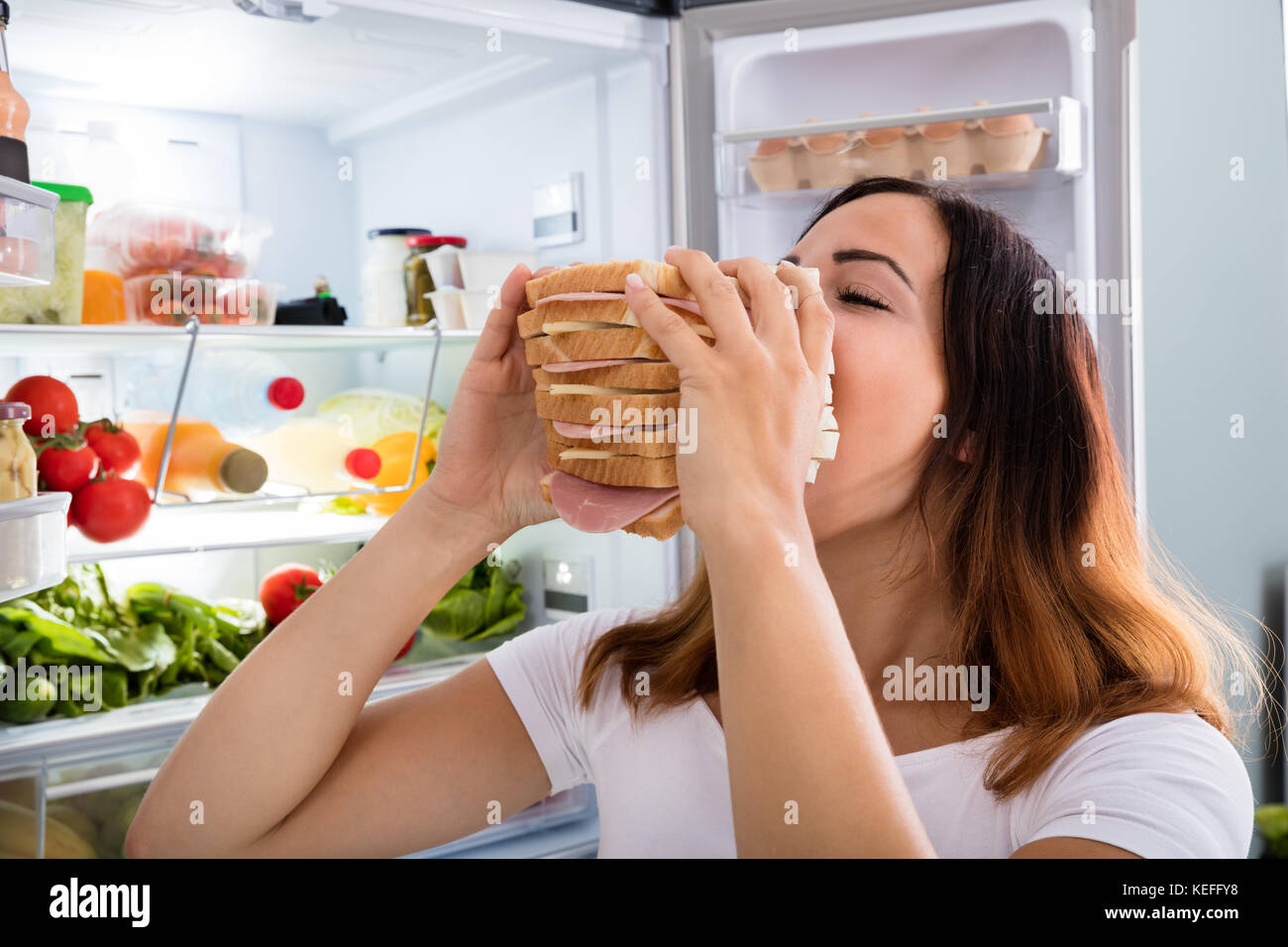 Young Woman Eating Meat Sandwich In Front Of Open Fridge Stock Photo