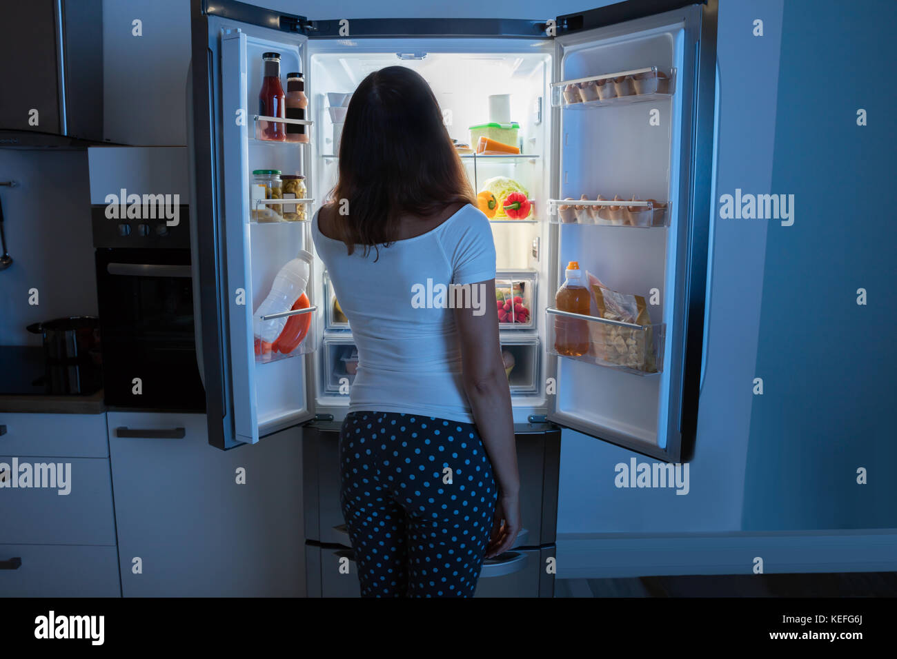 Rear View Of Young Woman Looking In Fridge At Kitchen Stock Photo