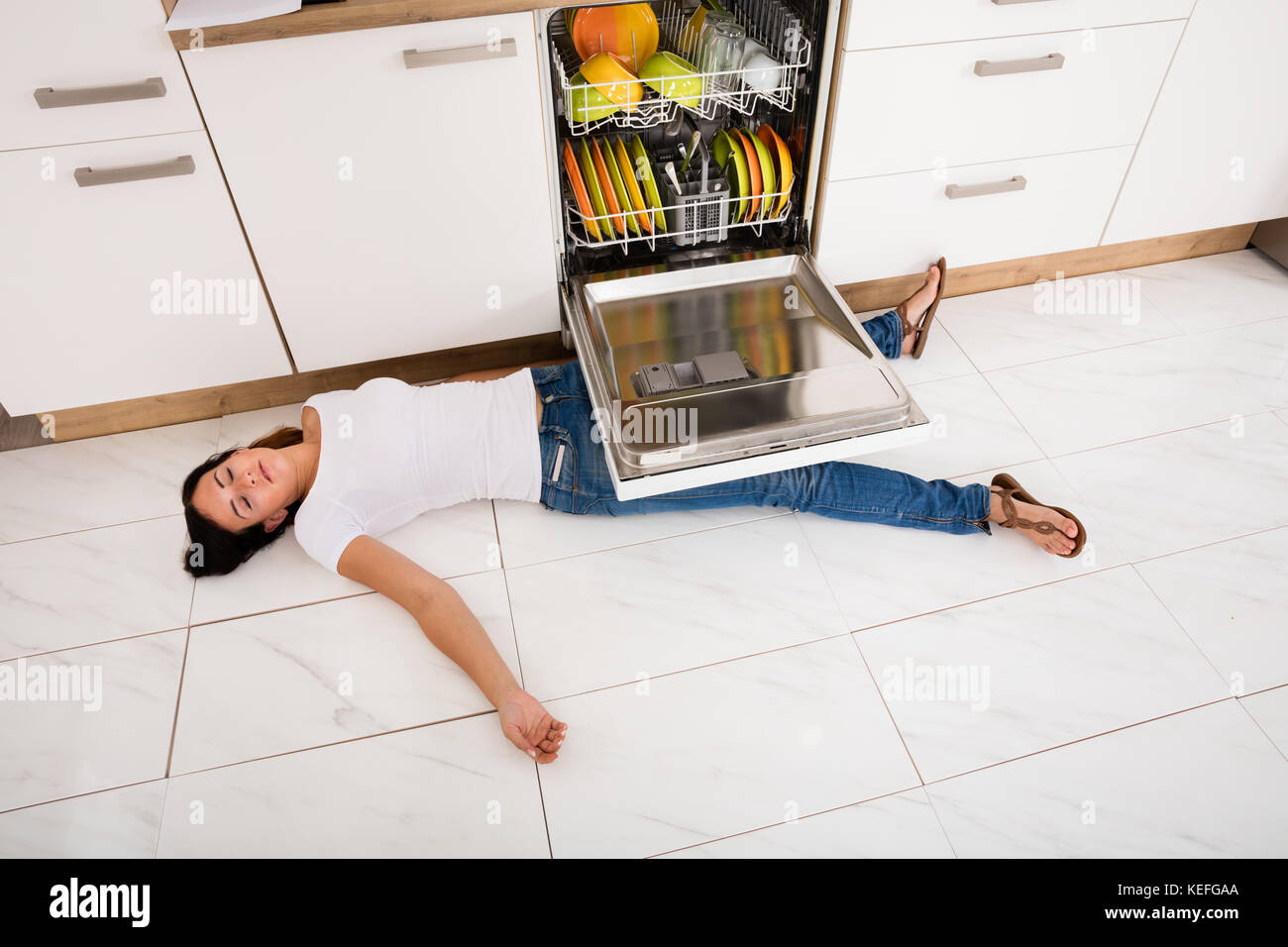 High Angle View Of Young Woman Lying On Floor Under A Dishwasher Stock