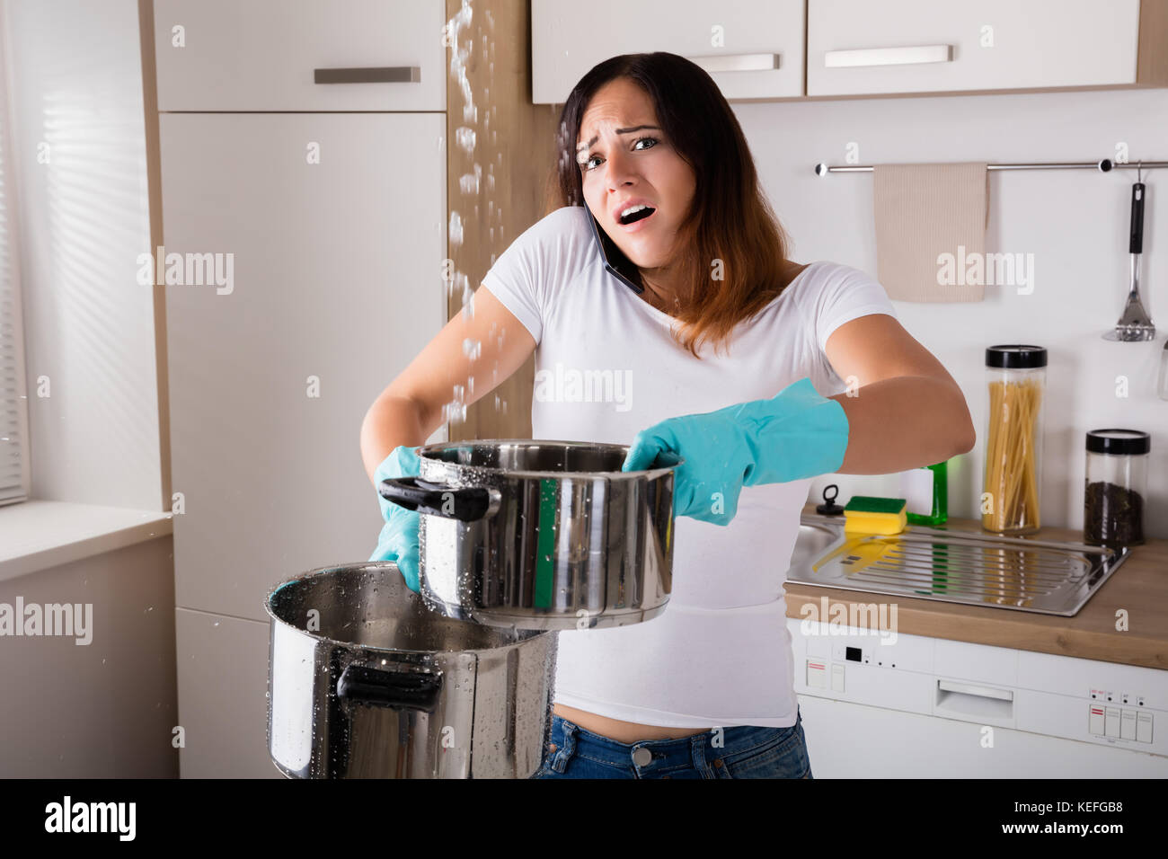 Shocked Woman Calling Plumber While Collecting Water Leaking From Ceiling Using Utensil Stock Photo