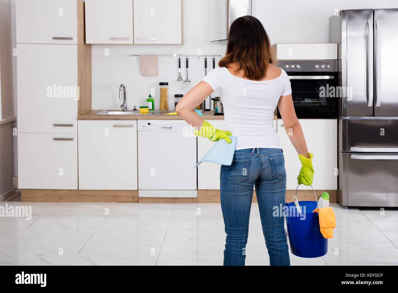 Rear View Of Woman Holding Cleaning Tools And Products In Bucket At Kitchen Stock Photo