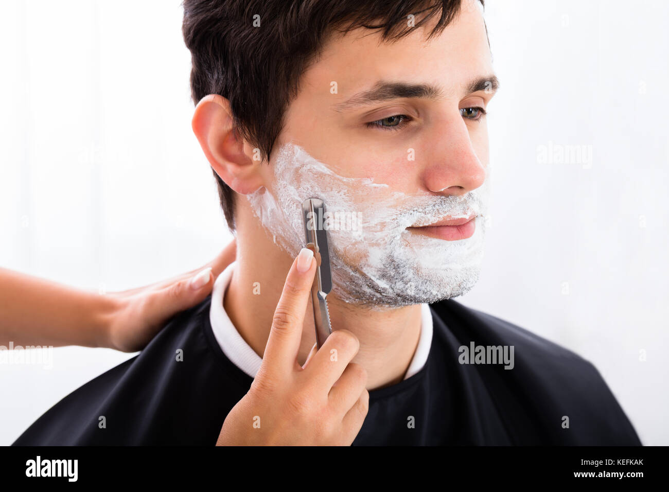 Hairdresser Shaving Young Man's Beard With Razor At Salon Stock Photo