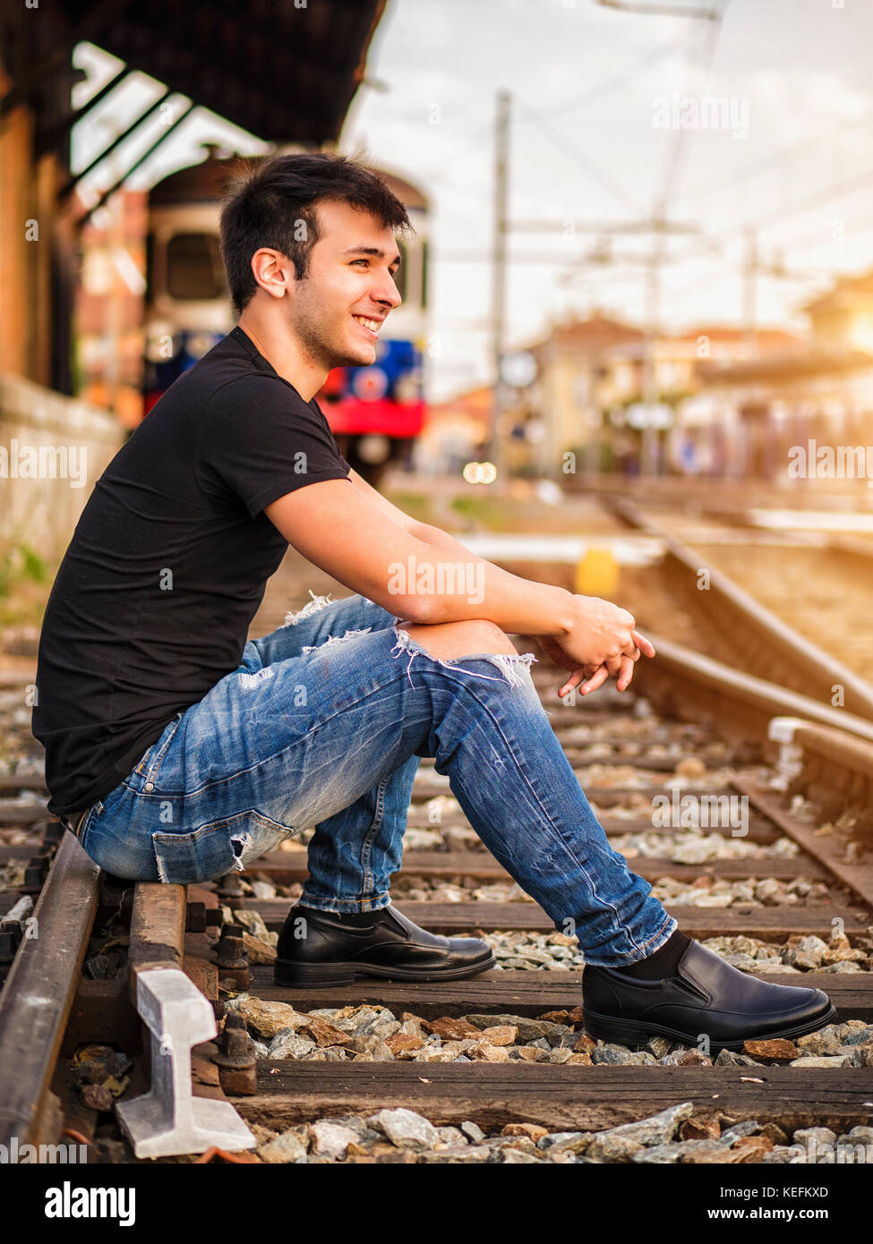 Attractive young man sitting on railroad, wearing black t-shirt and jeans,  looking afar Stock Photo - Alamy