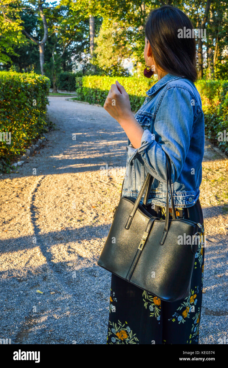 An outdoor portrait of a young woman, wearing a floral dress and a denim jacket, holding a black handbag Stock Photo