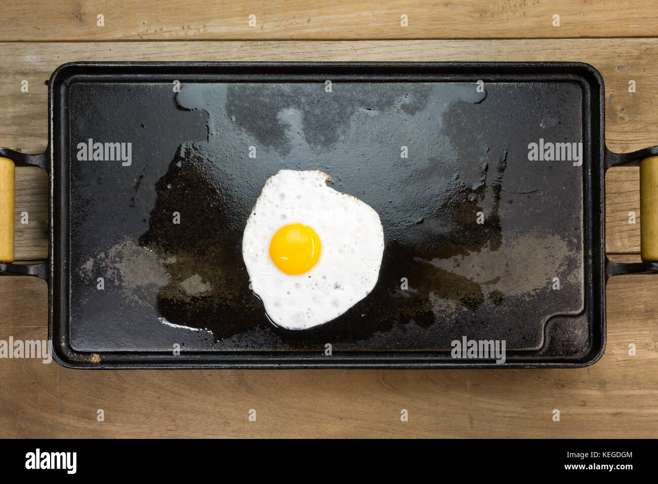 Fried egg white and yolk rectangular cast iron flat griddle pan with wooden handles, wood tabletop background, flat lay Stock Photo