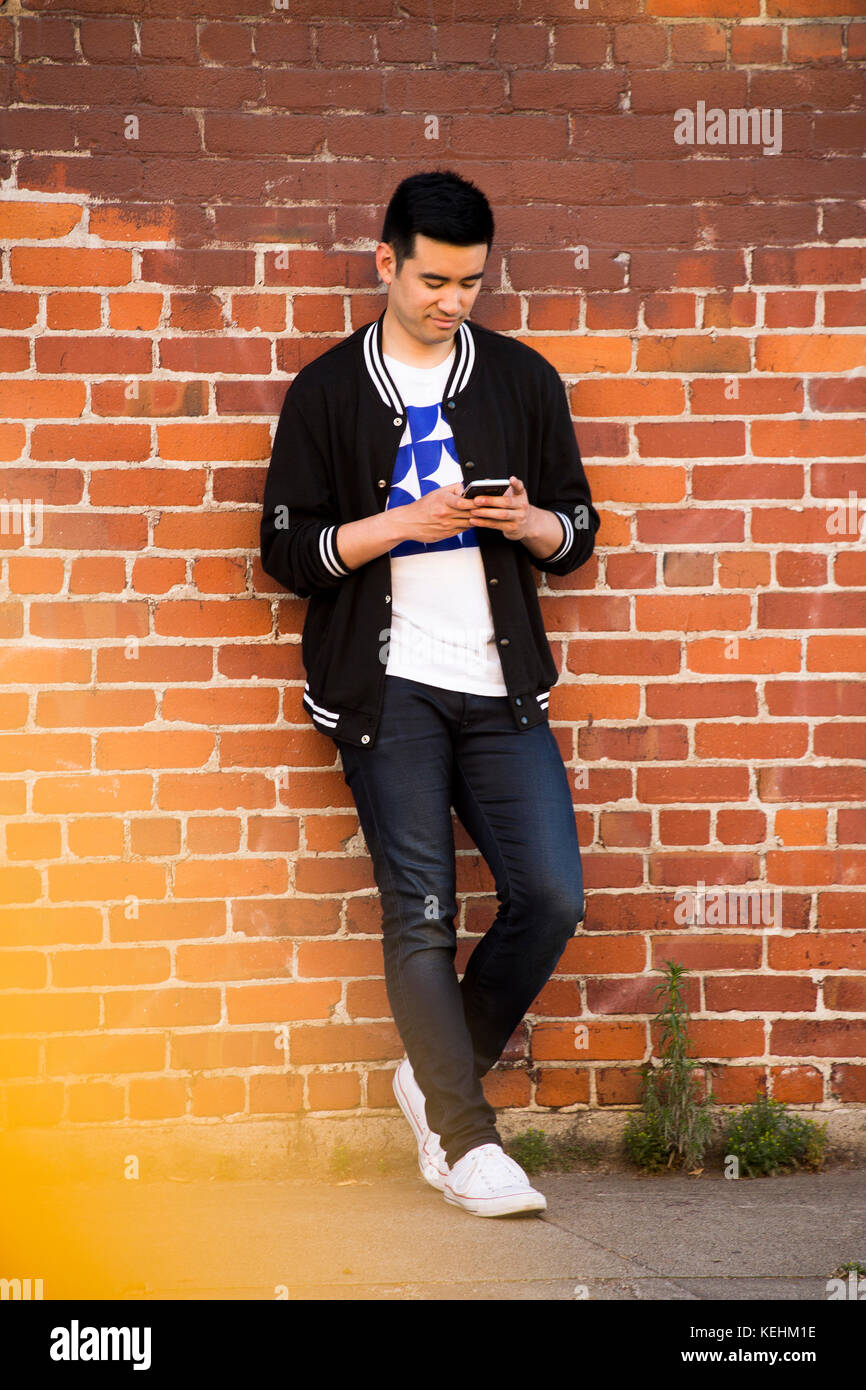 Chinese man leaning on brick wall texting on cell phone Stock Photo