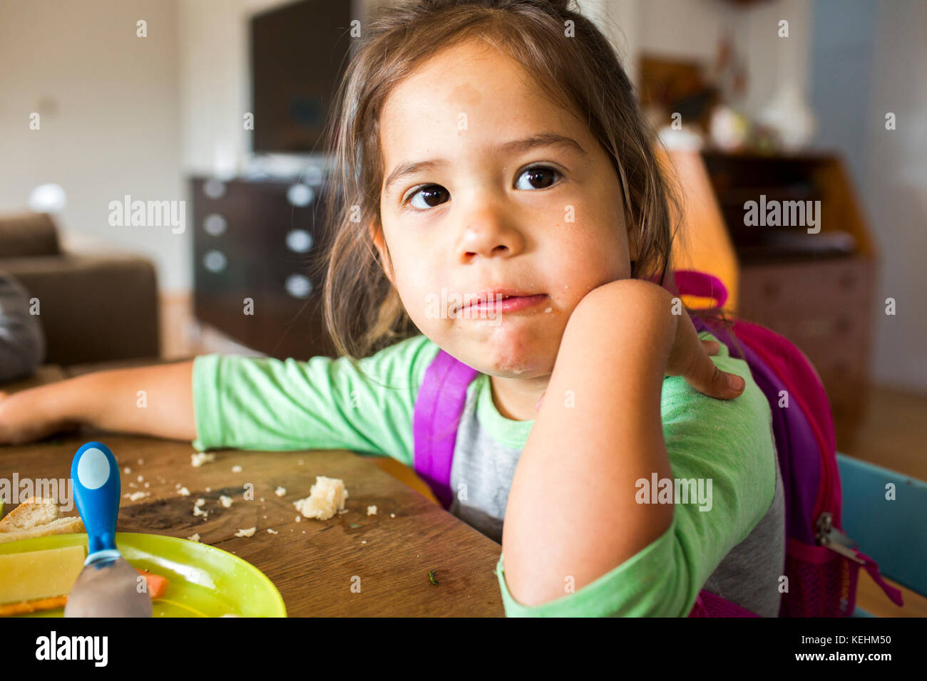Messy mixed race girl eating food and wearing backpack at table Stock Photo