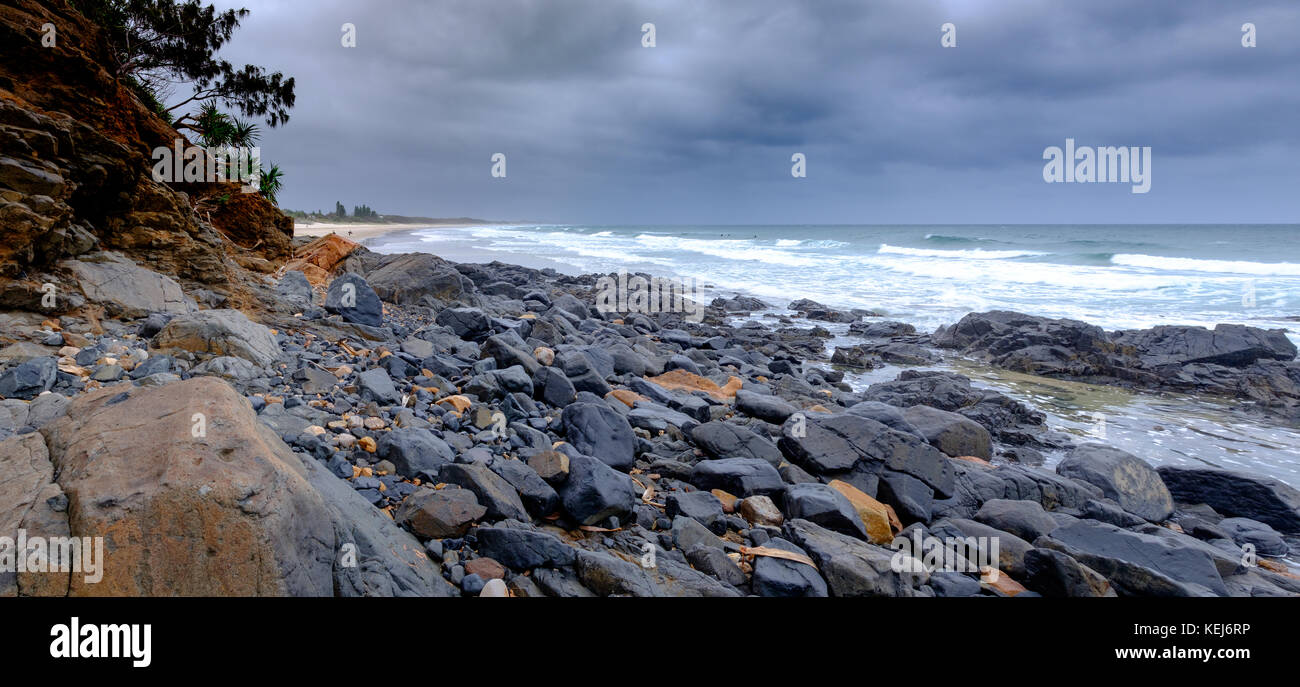 Rocky shore with stormy sky and surfers in the distance, Coolum Beach, Sunshine Coast, Queensland, Australia Stock Photo