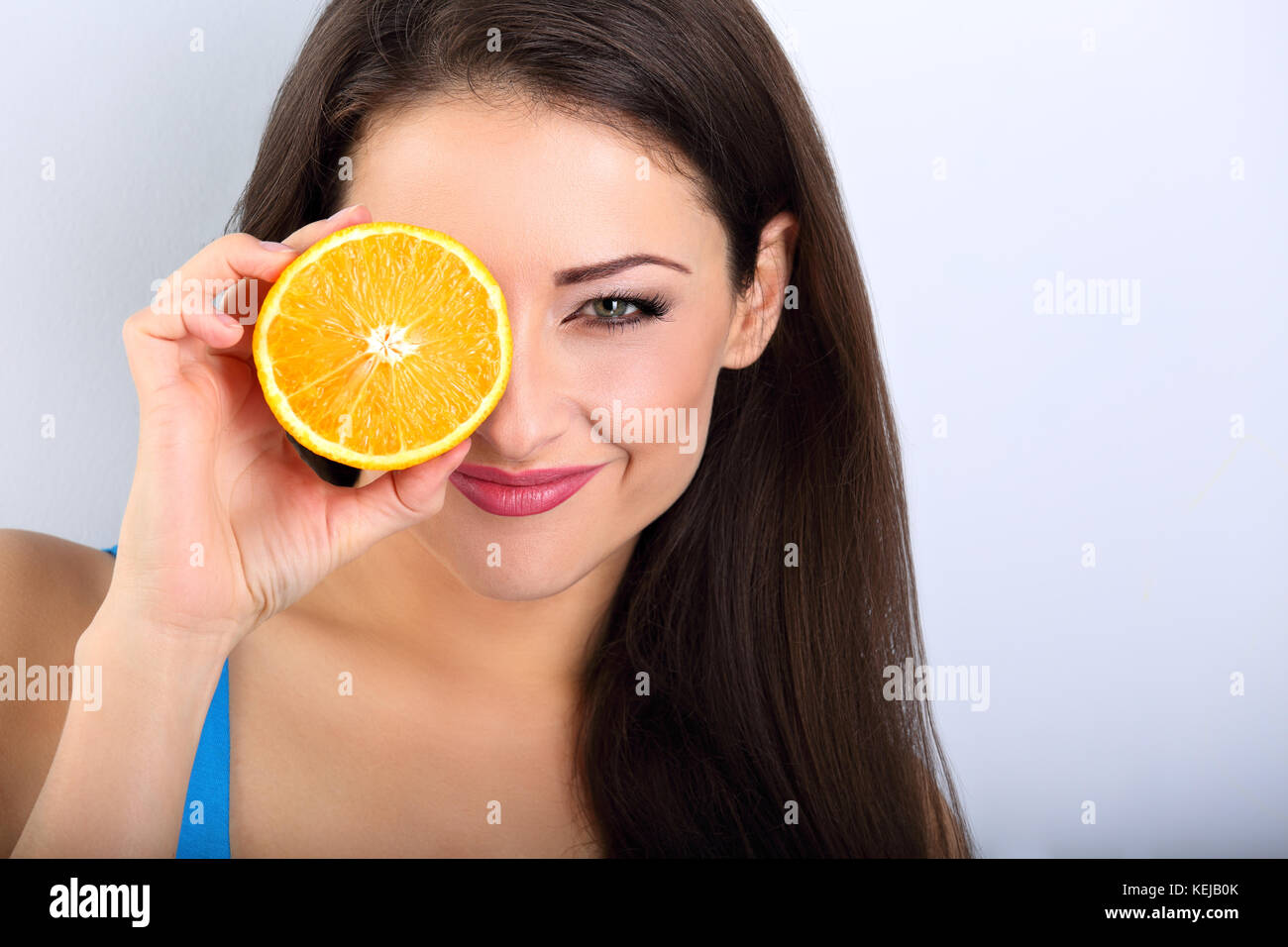 Beautiful makeup brunette woman holding fresh orange fruit near the eye and looking happy with empty space blue background. Closeup portrait Stock Photo
