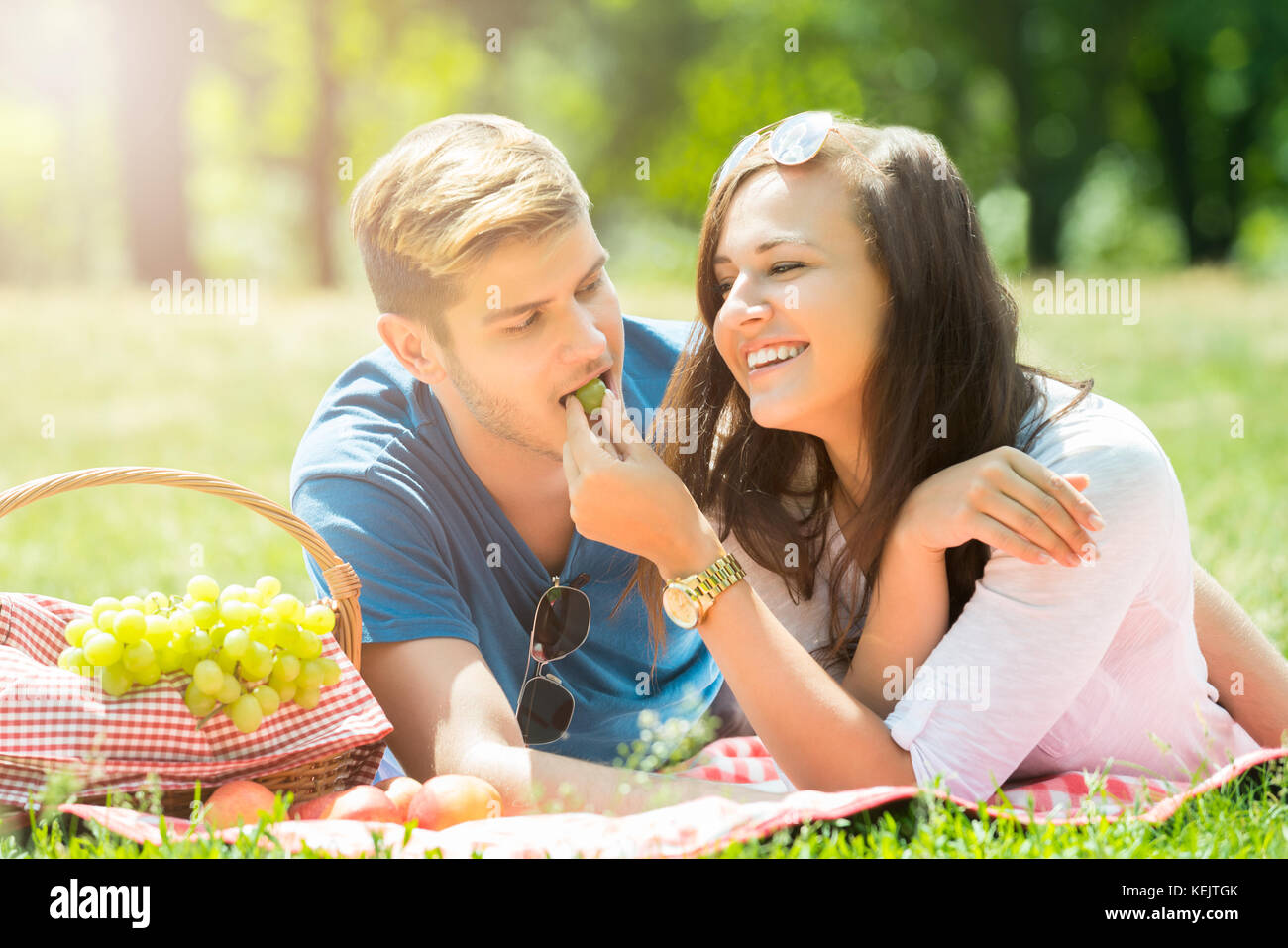 Smiling Young Woman Feeding Grape To Her Boyfriend In Park Stock Photo