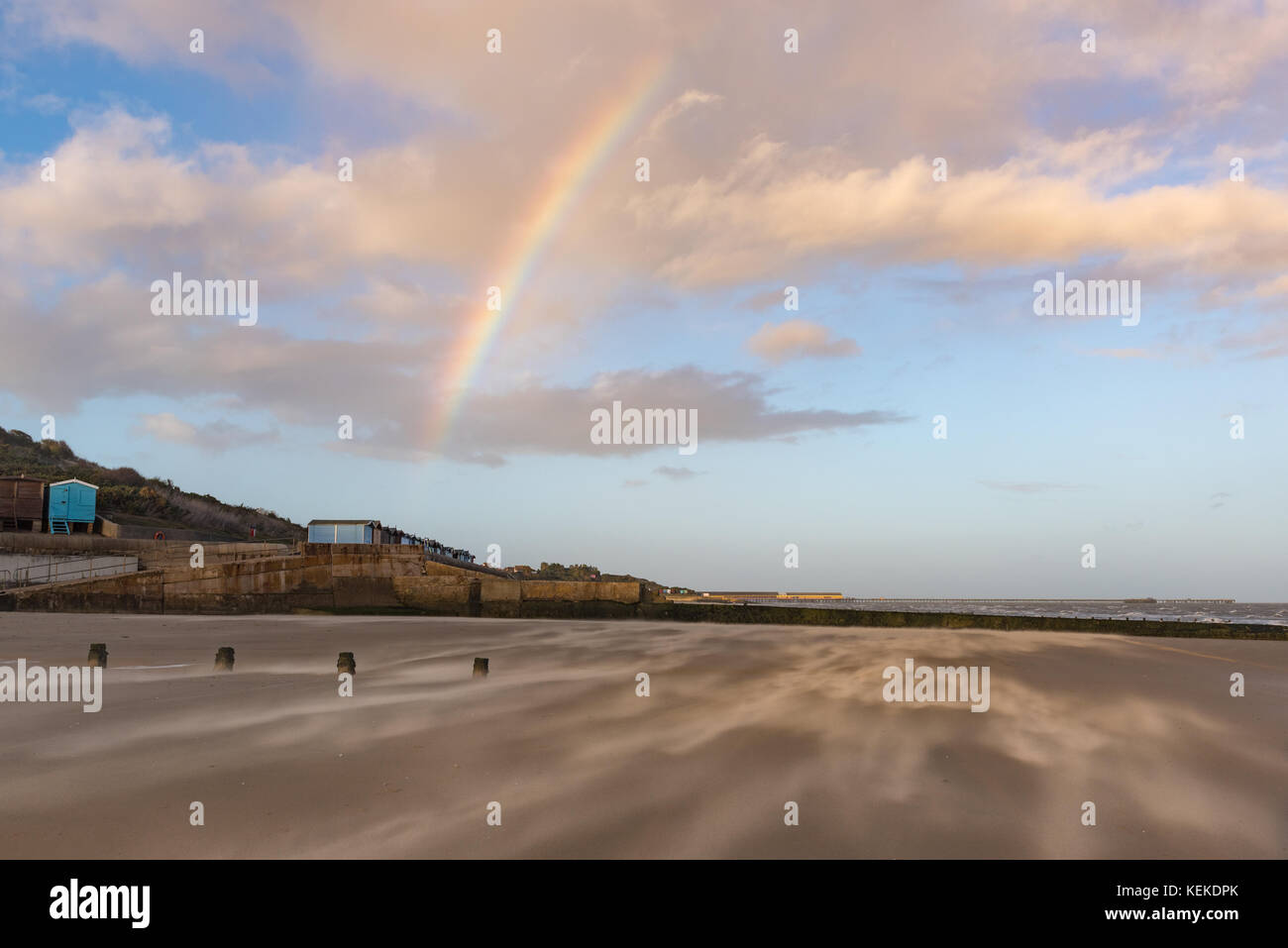 Frinton beach, Essex, UK. 21st October, 2017. At the end of our afternoon walk, the rising wind from Storm Brian was blasting the sand along the beach at Frinton-on-Sea in Essex. With the sun setting bright behind us, a rainbow appeared ahead in the most unlikely location. Looking north towards the pier at Walton. Credit: Gary Eason/Alamy Live News Stock Photo