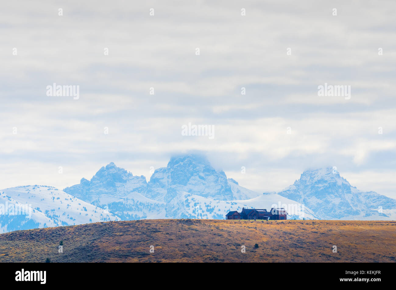 An Idaho home is lifted by the rolling hills and placed against the distant Grand Teton Mountains covered in a thick layer of velvety white clouds. Stock Photo