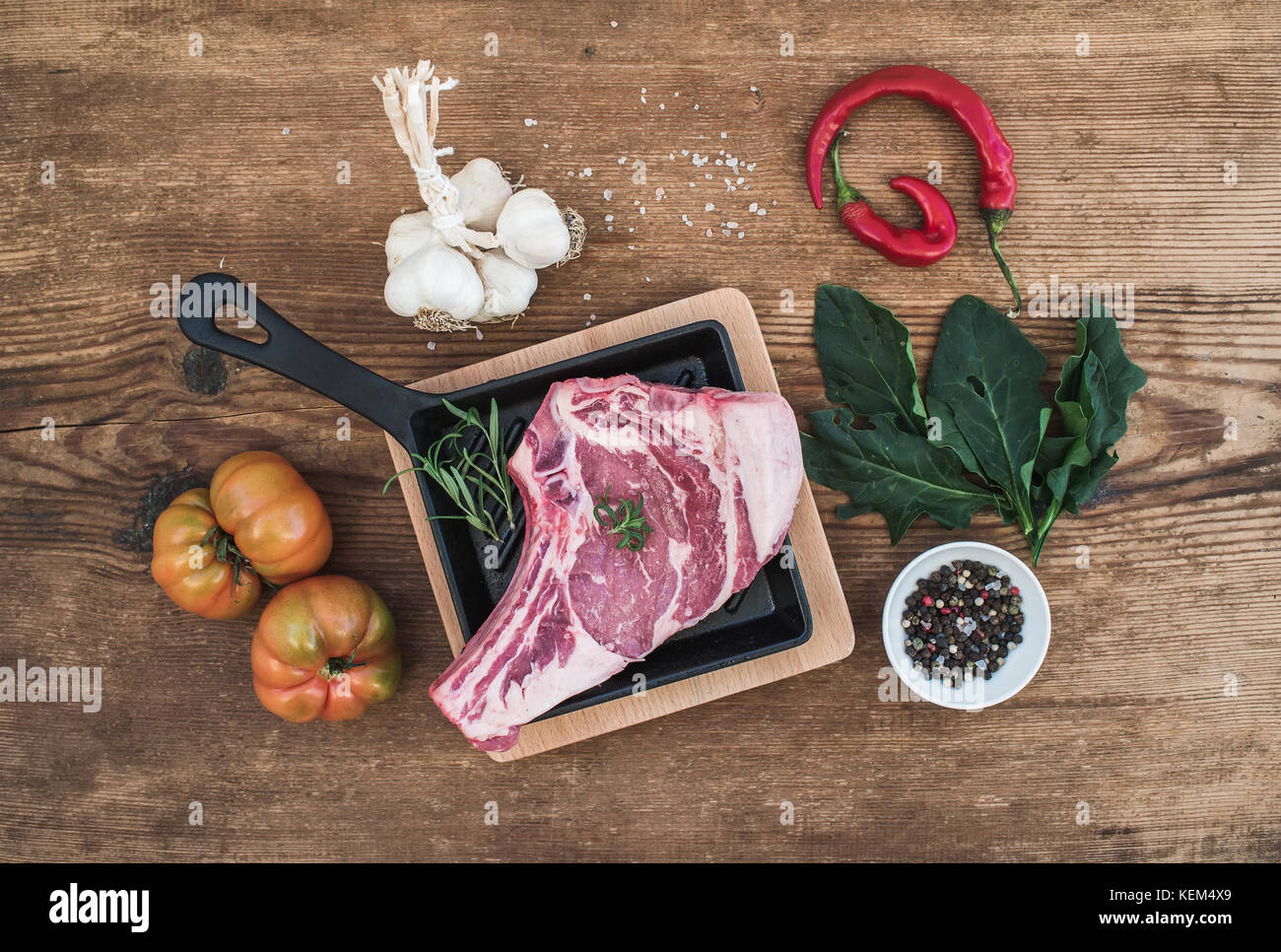Raw fresh meat ribeye steak with pepper, salt, chili, garlic, spinach, heirloom tomatoes and rosemary in cooking pan over rustic wooden background, top view. Stock Photo