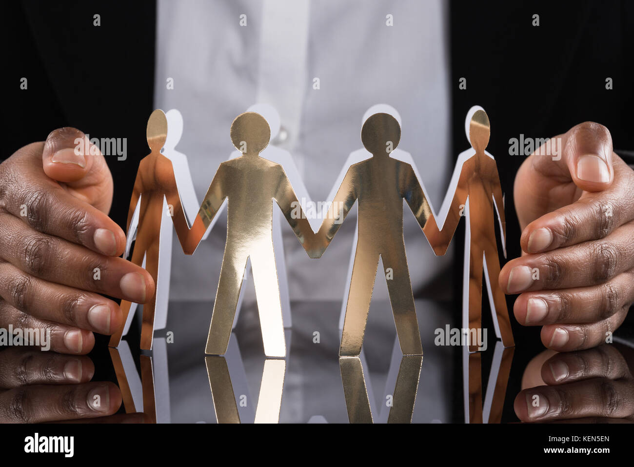 Close-up Of Businessperson Protecting Cut-out Figures On Desk Stock Photo