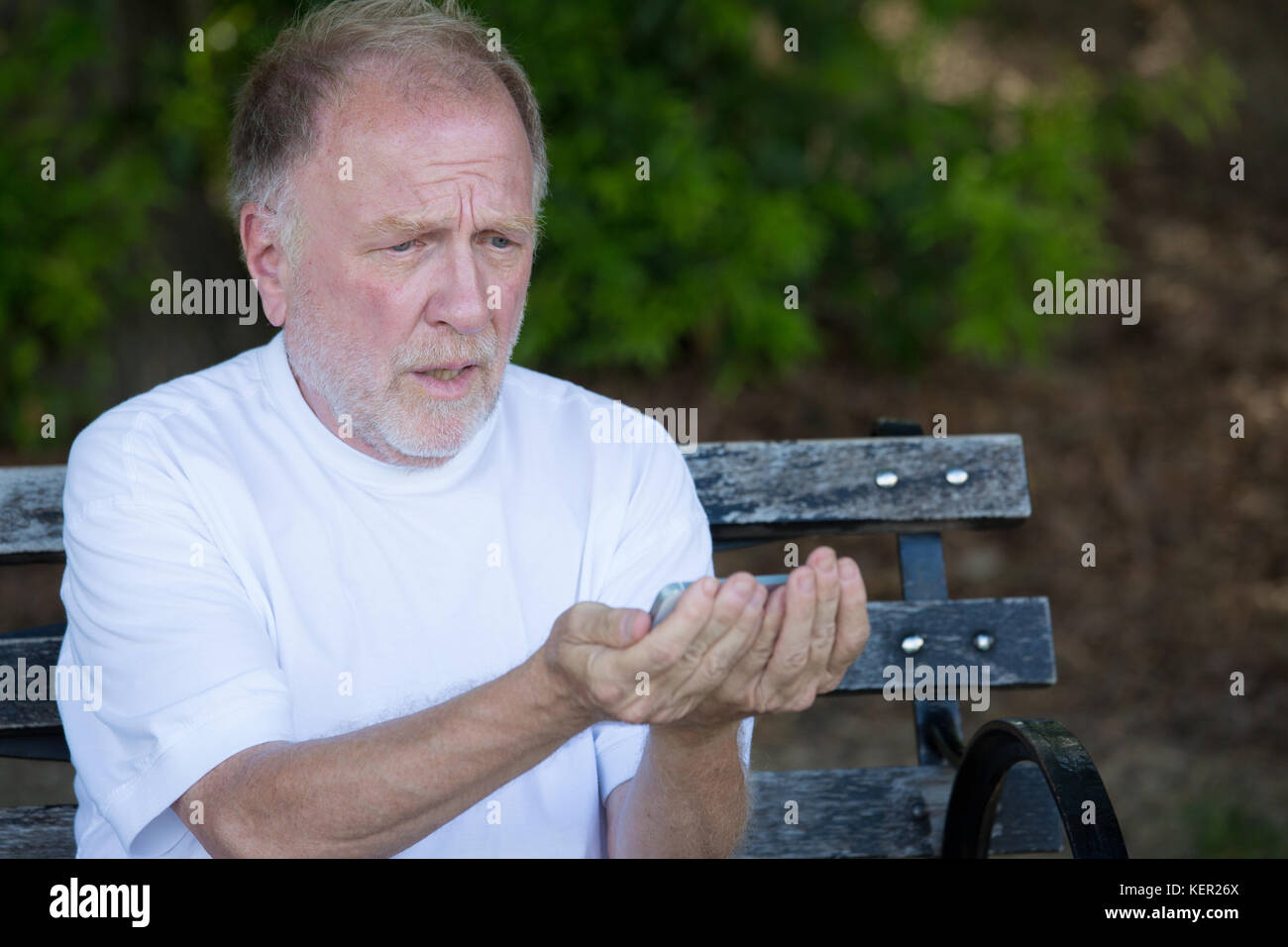 Closeup portrait of bold elderly man in white shirt, squinting, checking smartphone, sending text message, seated on a bench, isolated outdoor backgro Stock Photo