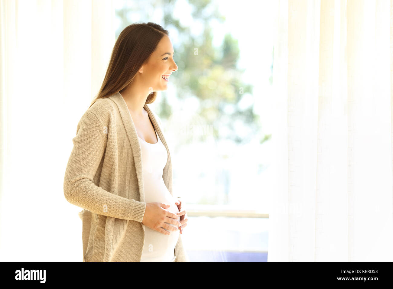 Side view portrait of a pregnant lady looking through a window standing at home Stock Photo