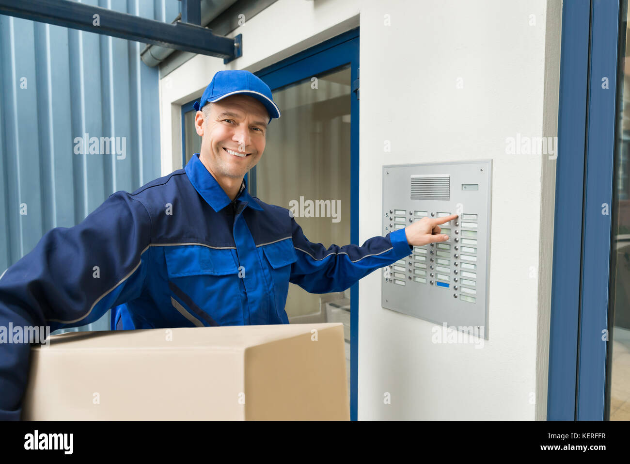 Delivery Man With Cardboard Box Pressing Button Of Intercom To Enter Building Stock Photo