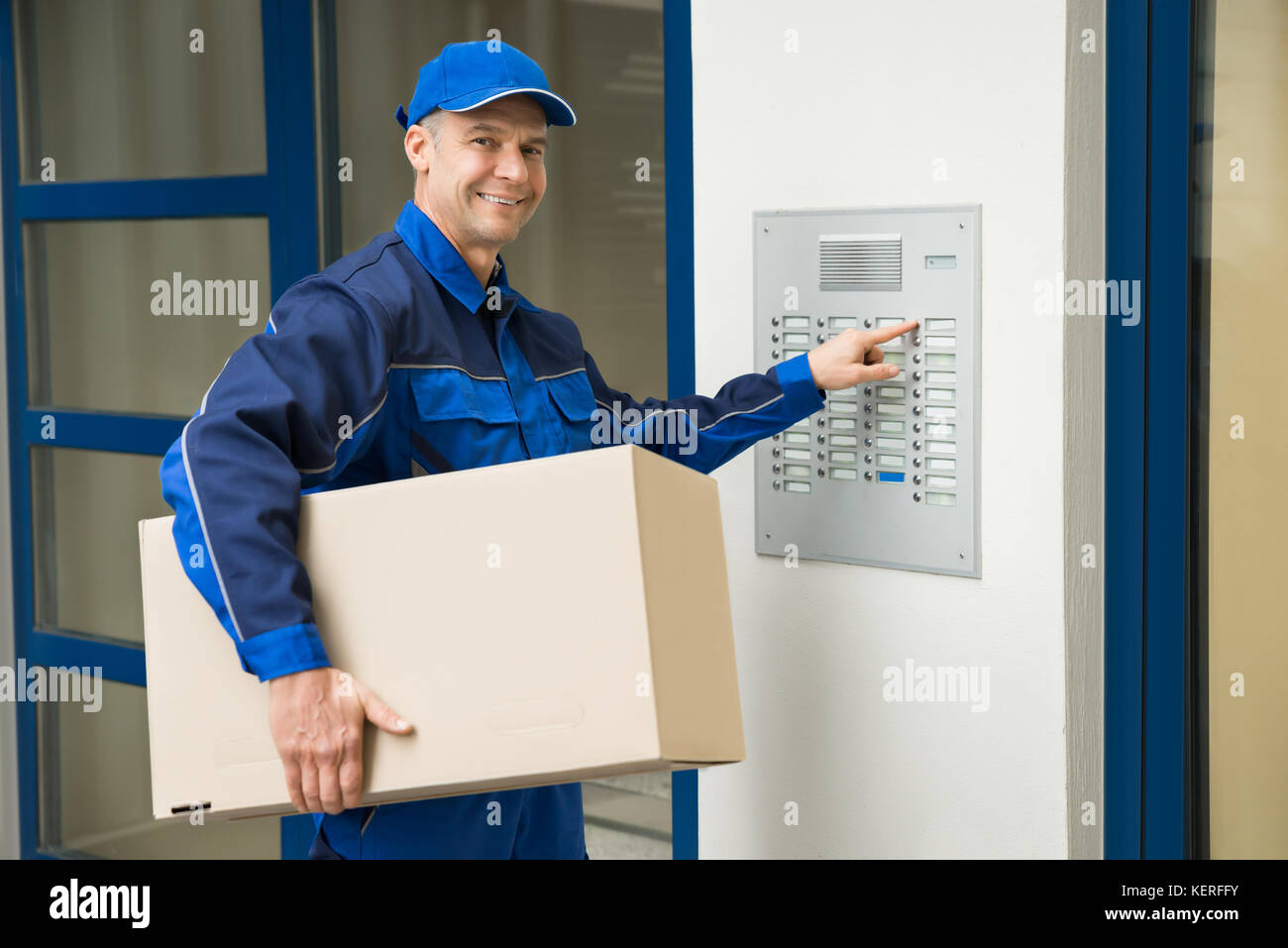 Delivery Man With Cardboard Box Pressing Button Of Intercom To Enter Building Stock Photo