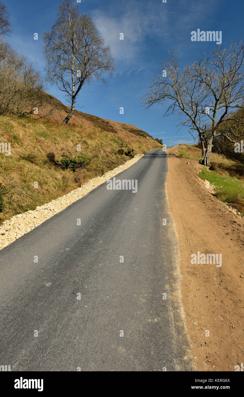 A narrow moorland road climbing a steep hillside prone to flood damage. A roadside drainage channel has been filled with crushed stone. Stock Photo