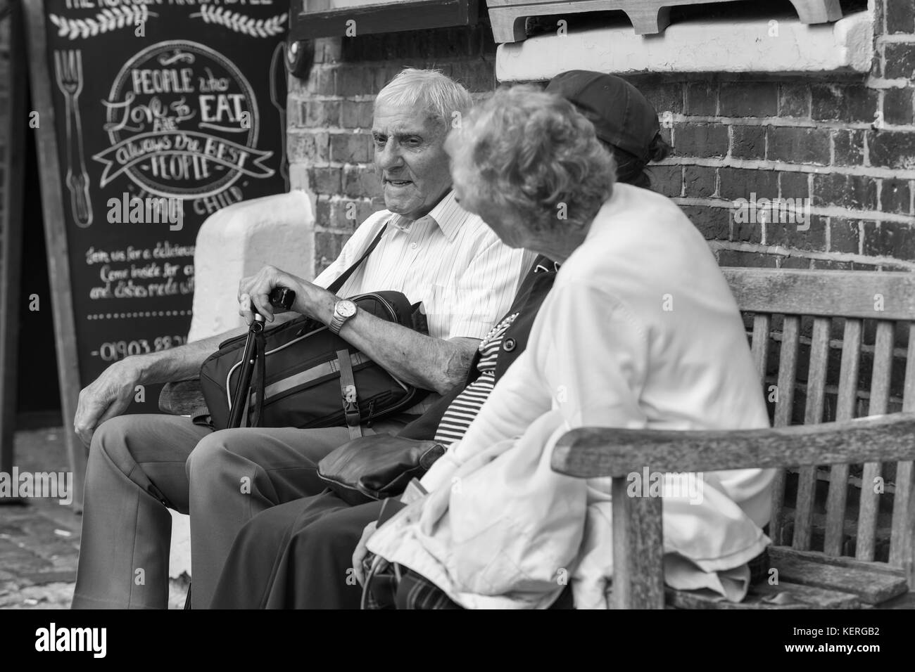 Elderly people sitting on a wooden bench outside catching up, in black and white monotone. Stock Photo
