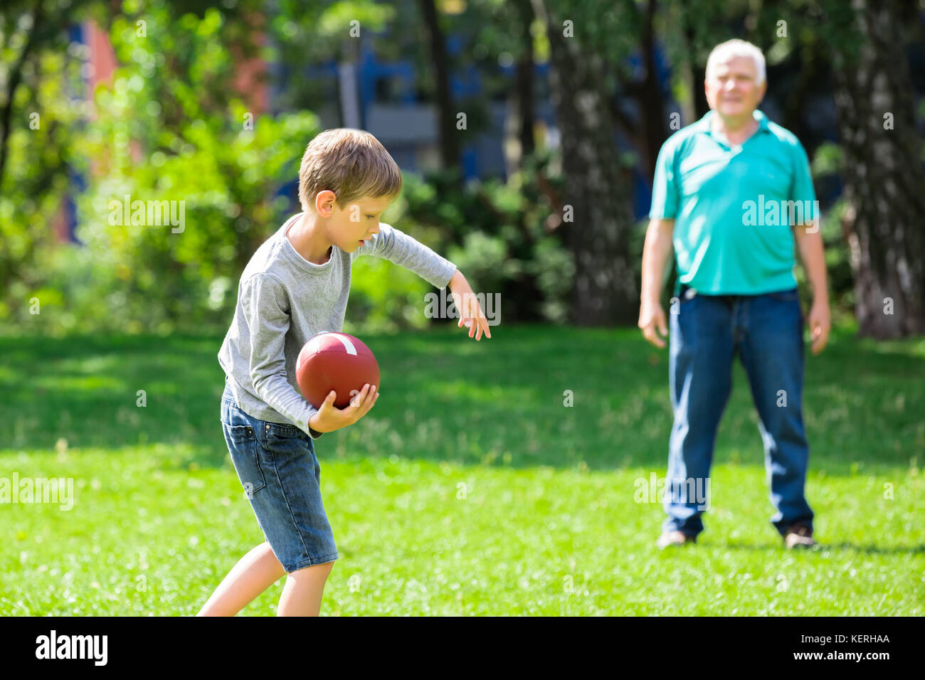 Kid Throwing Rugby Ball To Grandfather While Playing In The Park Stock Photo