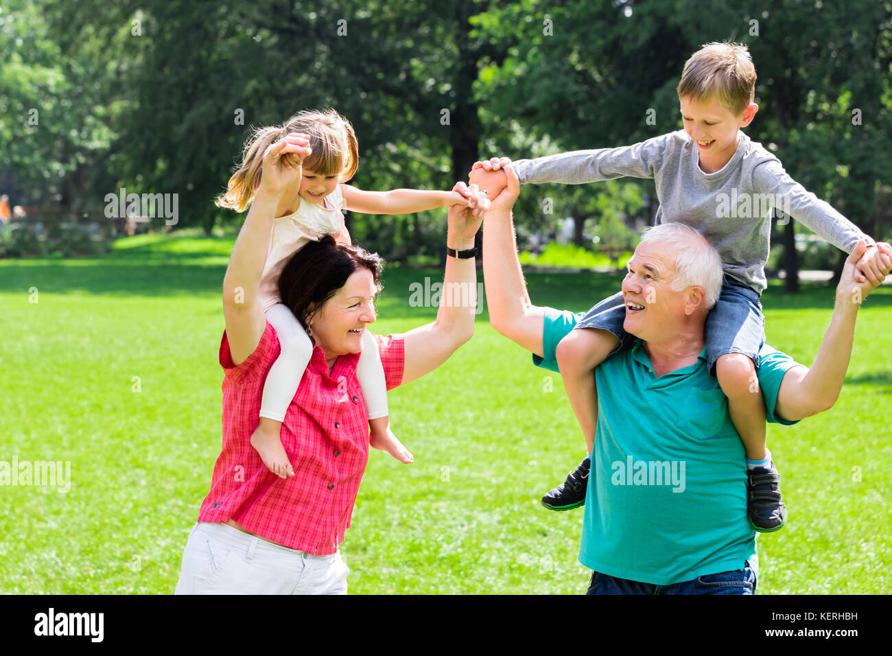 Happy Elderly Grandparents Enjoy Fun Piggyback Ride With Kids Together In Park Stock Photo