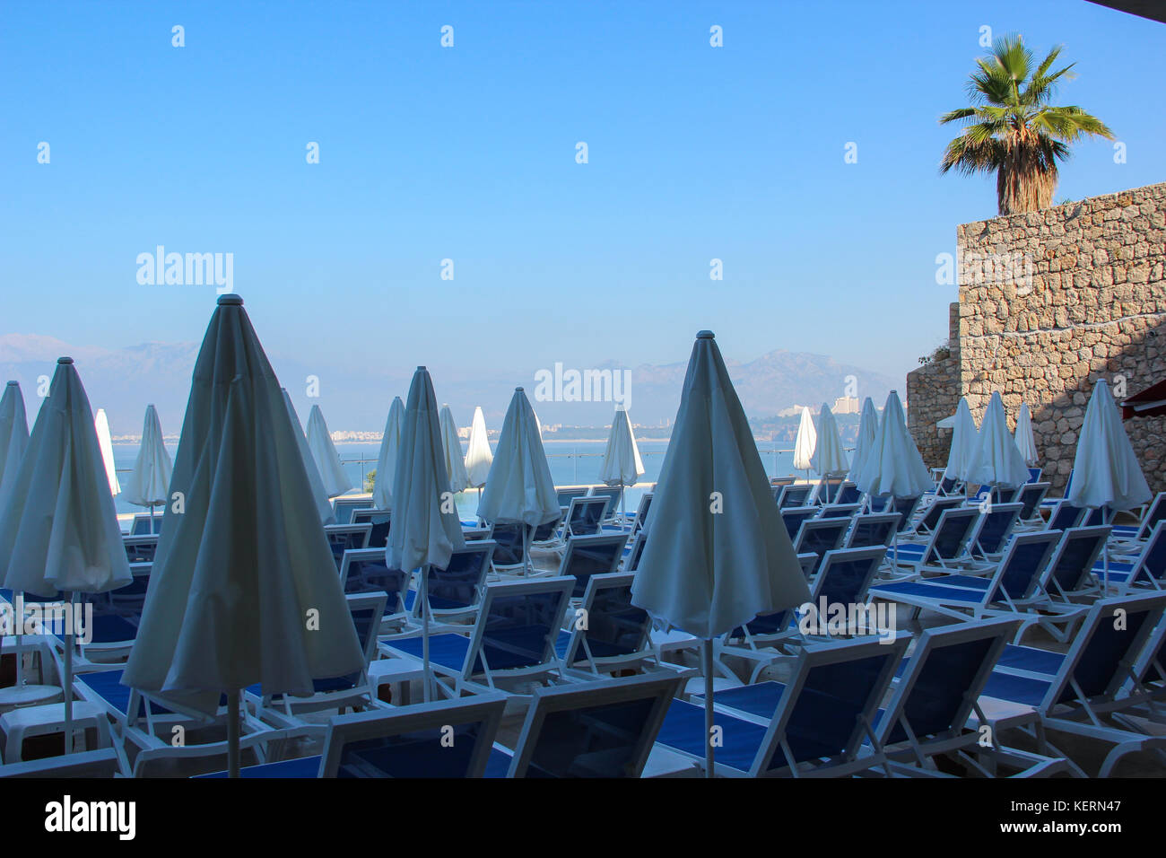 The view of the chairs and closed sunshades against the bright sky, the azure sea, the mountain range. Early morning of a Sunny summer day. travel Stock Photo