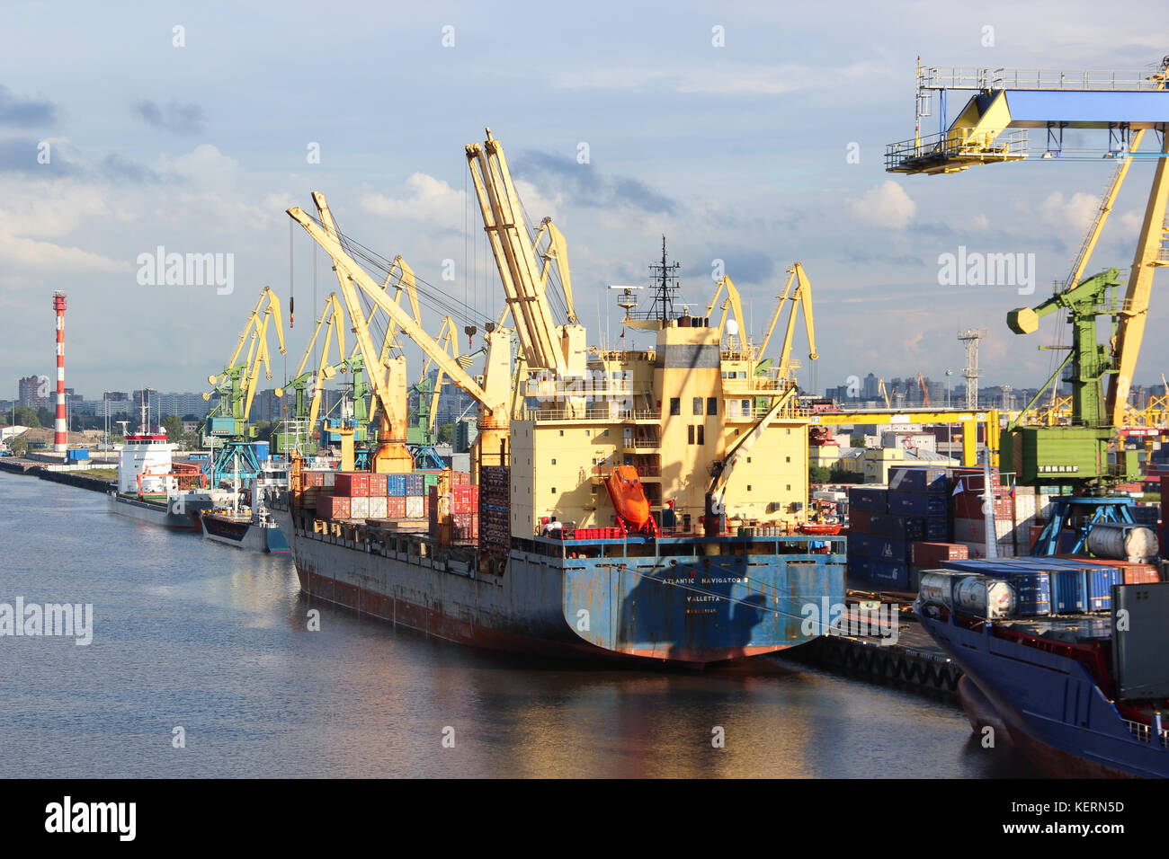 The cargo port. Containers with goods are loaded / unloaded on a commercial vessel using a crane in the harbor of the city. Summer. Business Stock Photo