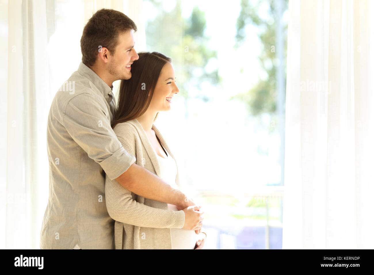 Side view portrait of a pregnant woman and husband looking through a window standing at home Stock Photo