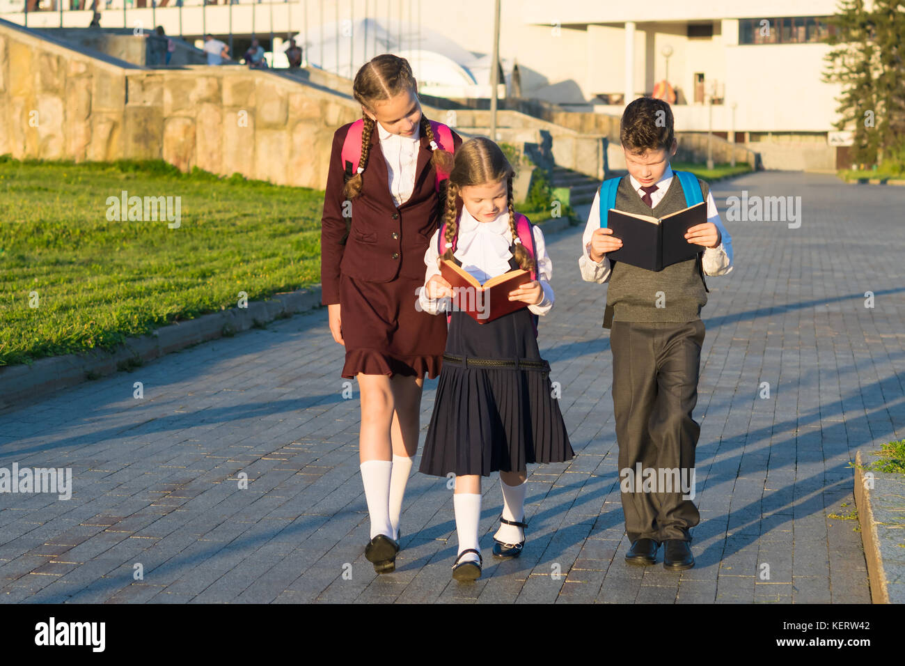 Three pupils in school uniform walking along the road in the park and reading a book Stock Photo