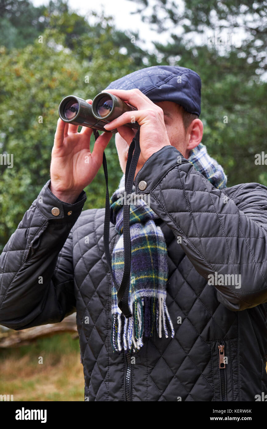 Young male dressed in winter clothing looking through binoculars in a woodland setting Stock Photo