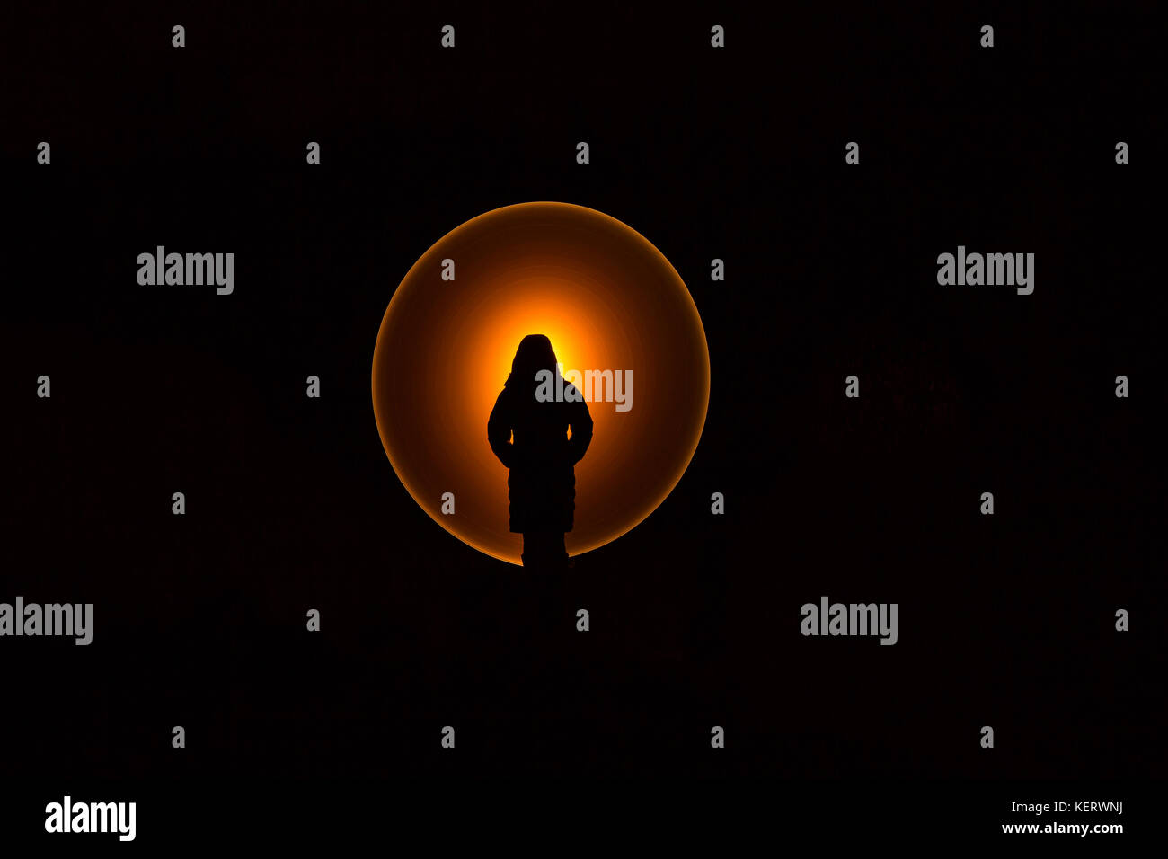Silhouette of figure, hands in pocket, against unexplained, mysterious circle of glowing, orange light. Blacked out night scene with special lighting. Stock Photo