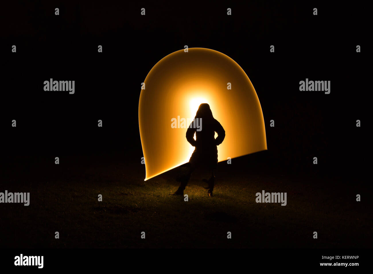 Silhouette figure of lone woman in coat standing isolated in the dark, black background with unexplained, golden light waves in arc. Light painting UK. Stock Photo
