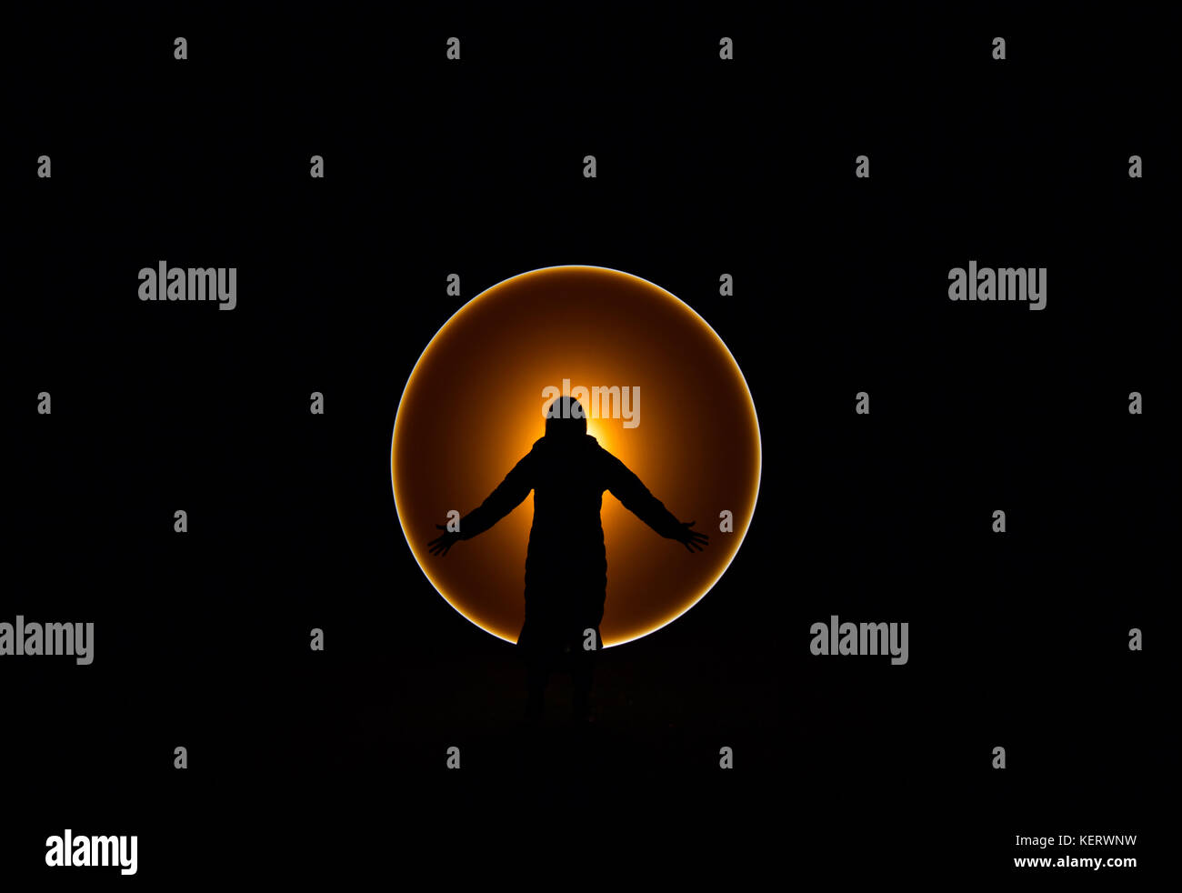 Silhouette of figure, arms outstretched, against unexplained, mysterious circle of glowing, orange light.Blacked out night scene with special lighting Stock Photo
