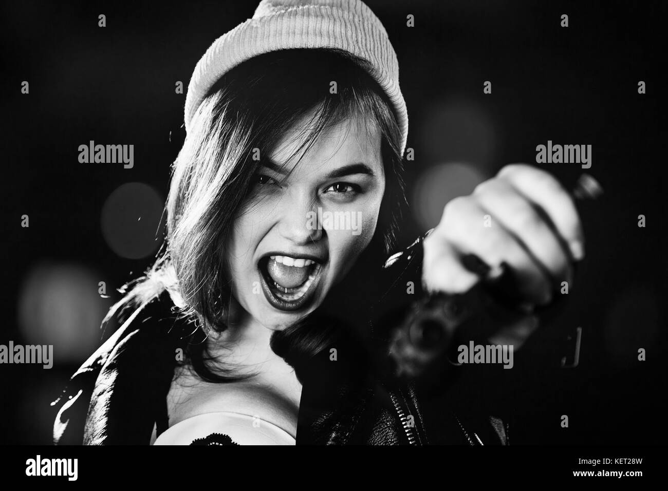 serious girl with gun on blurred background aiming at camera, screaming, monochrome Stock Photo